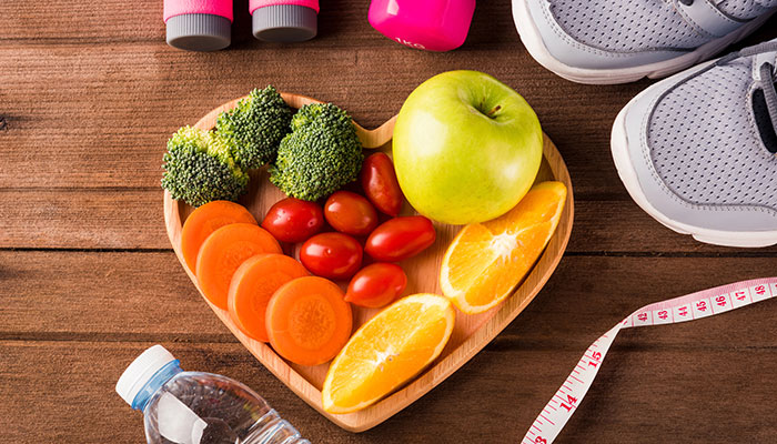 Draufsicht auf frisches Obst und Gemüse in Herzteller-Holz Apfel Karotte Tomate Orange Brokkoli und Hanteln Sportschuhe Sportgeräte auf Holztisch Ernährungskonzept Gesunder Lebensstil