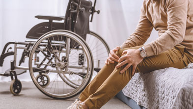 Mann sitzt auf Bett und hält sich die Beine, dahinter ein Rollstuhl, Copyright Panthermedia; Link zum Artikel Pflegeversicherung