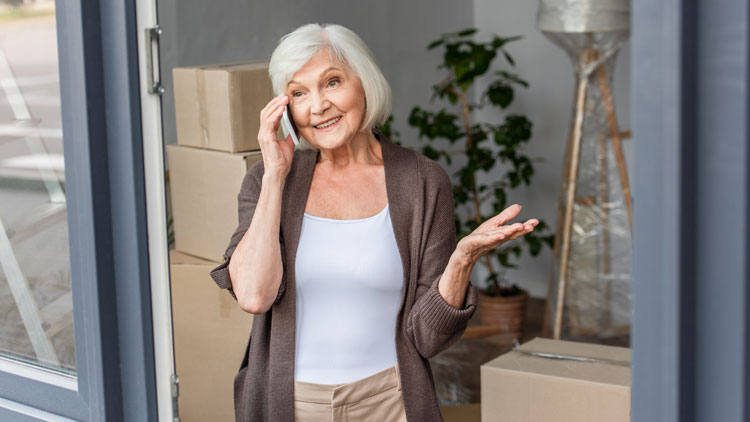 Grauhaarige Frau telefoniert vor Umzugskartons an der Terassentür einer Wohnung; Copyright Panthermedia